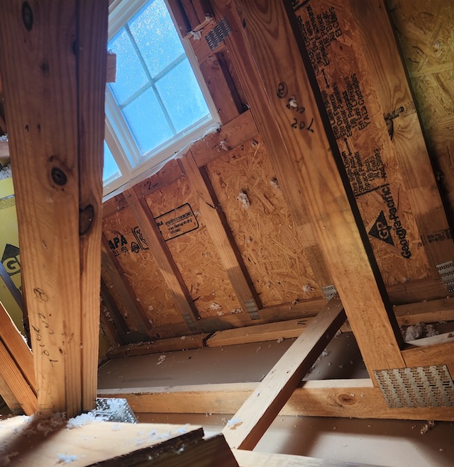 Interior view of a New Jersey attic showing exposed framing and roof sheathing near a frosted window, with scattered insulation indicating heat loss and ventilation issues.